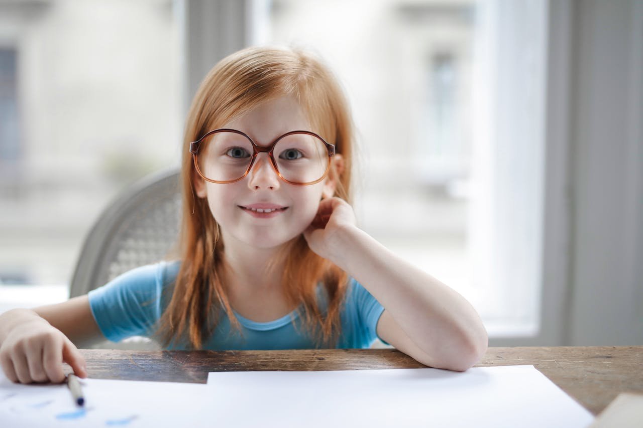 Smiling girl wearing eyeglasses, sitting indoors and doing homework at a table.